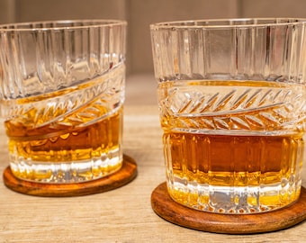May include: Two clear glass tumblers with intricate etched designs, filled with amber-colored liquid, resting on wooden coasters.