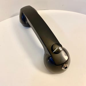 May include: A black vintage telephone handset with a glossy finish. The handset features a rounded earpiece and mouthpiece, connected by a rectangular bar. A small dial and a jack are visible on the handset.