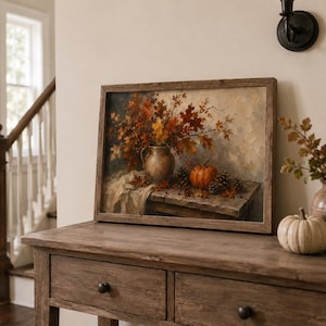 May include: A framed oil painting print featuring a rustic still life scene with a ceramic pitcher filled with autumn leaves, a small orange pumpkin, and pinecones resting on a wooden table, all rendered in warm brown, orange, and cream tones.