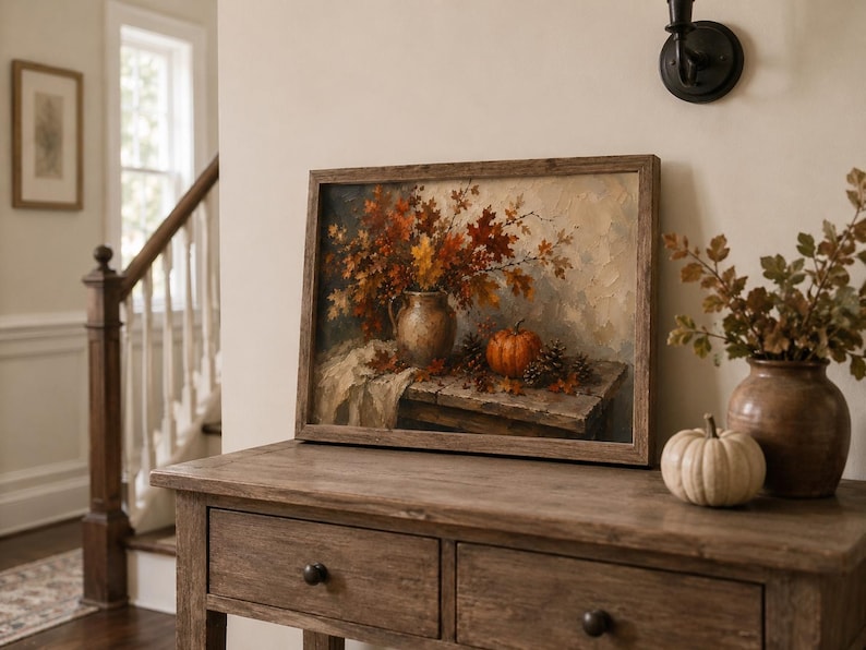 May include: A framed oil painting print featuring a rustic still life scene with a ceramic pitcher filled with autumn leaves, a small orange pumpkin, and pinecones resting on a wooden table, all rendered in warm brown, orange, and cream tones.