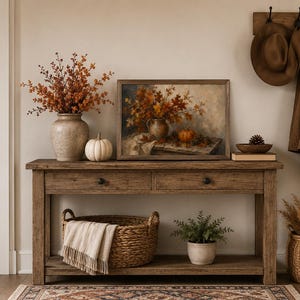 May include: A rustic wooden console table featuring a beige ceramic vase with orange autumn branches, a white pumpkin, and a framed painting of fall foliage. Below, a woven basket holds a cream throw, alongside a potted green plant.
