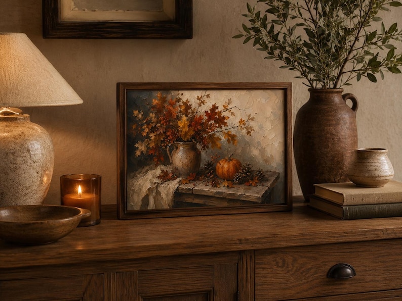 May include: A rectangular framed oil painting depicting a still life scene with a ceramic pitcher filled with autumn leaves, a small orange pumpkin, and pinecones resting on a wooden table draped with a piece of white fabric.
