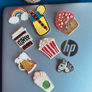 Peut inclure: Une collection de patchs thermocollants en perles colorées représentant différents motifs, dont un arc-en-ciel, une limonade, un cupcake, un gobelet à café, une chope de bière, un seau de pop-corn, une licorne et un gobelet vert et blanc.