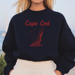 Peut inclure: Sweat-shirt bleu marine avec l'inscription "Cape Cod" en rouge et un dessin de voilier rouge. Le sweat-shirt est porté avec un pantalon blanc. Des voiliers sont visibles en arrière-plan.