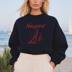 Puede incluir: Sudadera azul marino con la palabra "Newport" en rojo y un diseño de velero rojo. La sudadera se combina con pantalones blancos. Se ven veleros en el fondo.