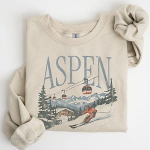 May include: A beige sweatshirt with a vintage-style graphic of a skier in Aspen, Colorado. The graphic includes a ski lift, mountains, and a cabin. The text "ASPEN" is printed in blue above the graphic.