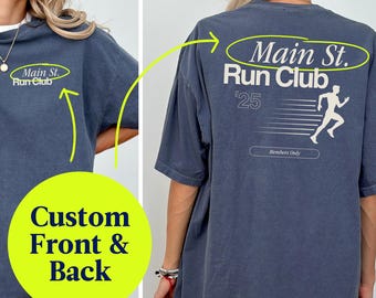 May include: A slate blue t-shirt with the text "Main St. Run Club" on the front and back. The back of the shirt features a running figure and the text "'25". The shirt has a relaxed fit.