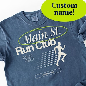 Może przedstawiać: Niebieska koszulka z napisem "Main St. Run Club" i grafiką biegacza. Koszulka zawiera również napis "'25" i "Members Only". Zielony owal z napisem "Custom name!" znajduje się na górze.