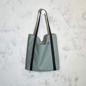 May include: A light green canvas tote bag with black straps. The bag has a single pocket on the front.