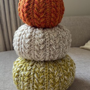 May include: A stack of three crocheted pumpkins in orange, cream, and olive green. Each pumpkin features a textured, ribbed stitch pattern. The top pumpkin is topped with a brown cinnamon stick stem, creating a vertical decorative arrangement.