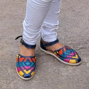 May include: A pair of colorful woven sandals with a blue base and yellow, red, and green accents. The sandals are worn on a person's feet with white jeans.