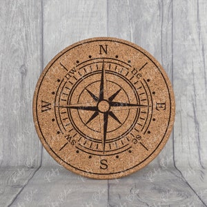 May include: A round cork coaster with a compass design. The compass features a black star with four points and the letters N, S, E, and W around the edge.