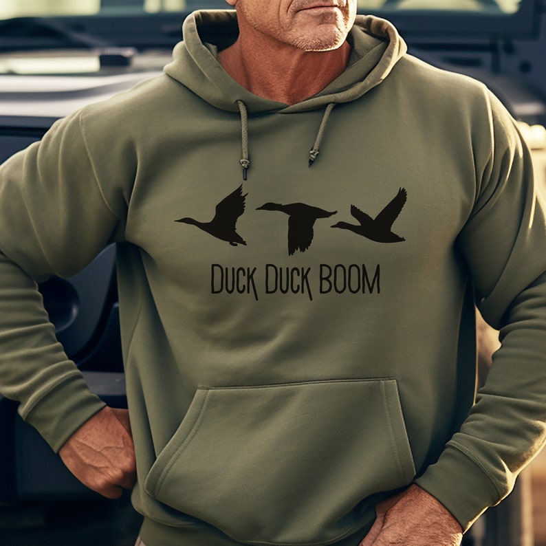 May include: A green hooded sweatshirt with a black graphic of three ducks flying in a row. The text "Duck Duck Boom" is printed below the ducks.