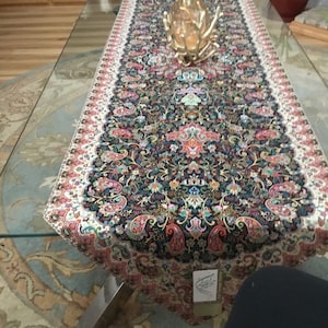 Puede incluir: Un camino de mesa negro y blanco con un patrón de paisley y acentos florales coloridos. El camino de mesa tiene un borde blanco con un borde festoneado.