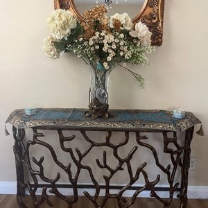 Puede incluir: Una consola decorativa con un marco de metal color bronce en forma de ramas. La mesa tiene un camino de mesa con estampado azul y dorado, y un jarrón de cristal transparente lleno de flores. Un espejo con marco dorado cuelga encima.