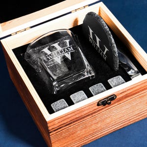 May include: A wooden gift box containing a personalised whiskey glass, a slate coaster, and whiskey stones. The glass and coaster are engraved with the name "WILLIAM". The box is lined with black fabric and has a metal clasp.