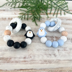 May include: Two baby teething rings. One features black, white, and wood-toned beads with a black penguin charm and a marble-patterned ring. The other has blue, white, and marble-patterned beads with a blue penguin charm.