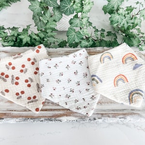 May include: Three white muslin baby bibs with different patterns. One bib has a red cherry pattern, one has a small floral pattern, and one has a rainbow pattern.