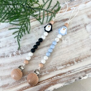 May include: Two baby pacifier clips with penguin designs. One clip features a black penguin, black beads, and white speckled beads. The other clip has a blue penguin, blue beads, and white speckled beads. Both have wooden clips and beads.
