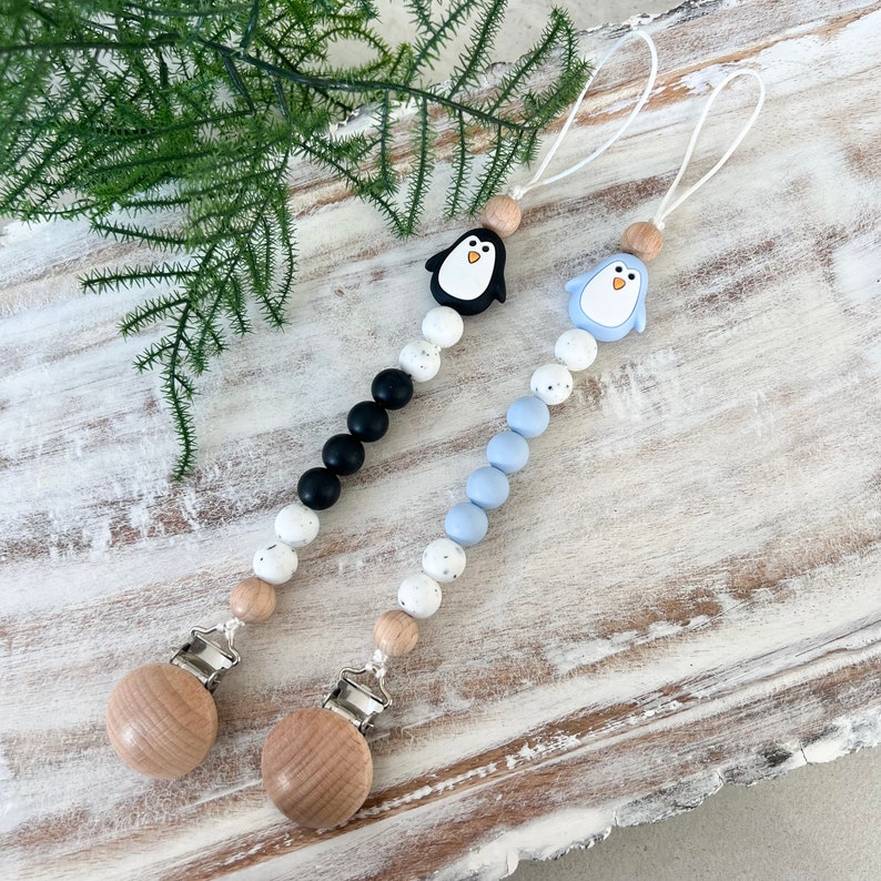May include: Two baby pacifier clips with penguin designs. One clip features a black penguin, black beads, and white speckled beads. The other clip has a blue penguin, blue beads, and white speckled beads. Both have wooden clips and beads.