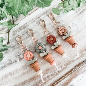 May include: Four keychains with gold-tone metal rings and a white cord. Each keychain features a small potted plant with a flower in shades of green, pink, and orange.