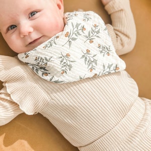K&ouml;nnte beinhalten: Ein Baby in einem cremefarbenen Outfit und einem L&auml;tzchen. Das L&auml;tzchen ist wei&szlig; mit einem Muster aus gr&uuml;nen Bl&auml;ttern und orangefarbenen Beeren. Das Outfit ist gerippt und hat R&uuml;schen&auml;rmel. Das Baby l&auml;chelt.