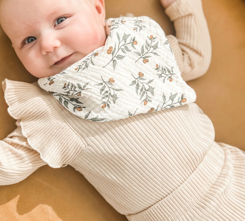 K&ouml;nnte beinhalten: Ein Baby in einem cremefarbenen Outfit und einem L&auml;tzchen. Das L&auml;tzchen ist wei&szlig; mit einem Muster aus gr&uuml;nen Bl&auml;ttern und orangefarbenen Beeren. Das Outfit ist gerippt und hat R&uuml;schen&auml;rmel. Das Baby l&auml;chelt.