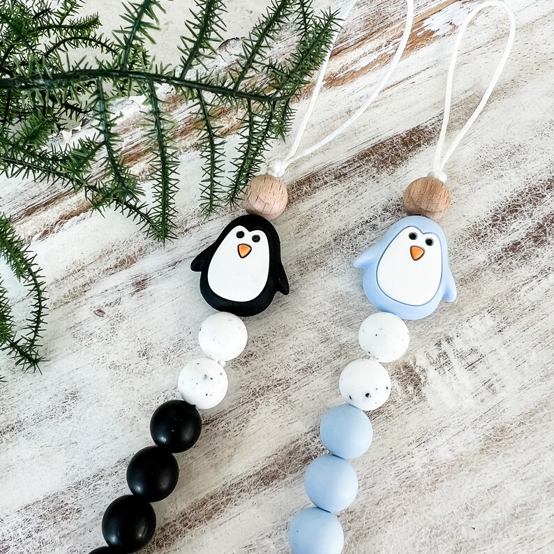 May include: Two penguin-themed pacifier clips. One clip features a black penguin with a white belly and orange beak, while the other has a light blue penguin. Both have wooden beads and white cords.