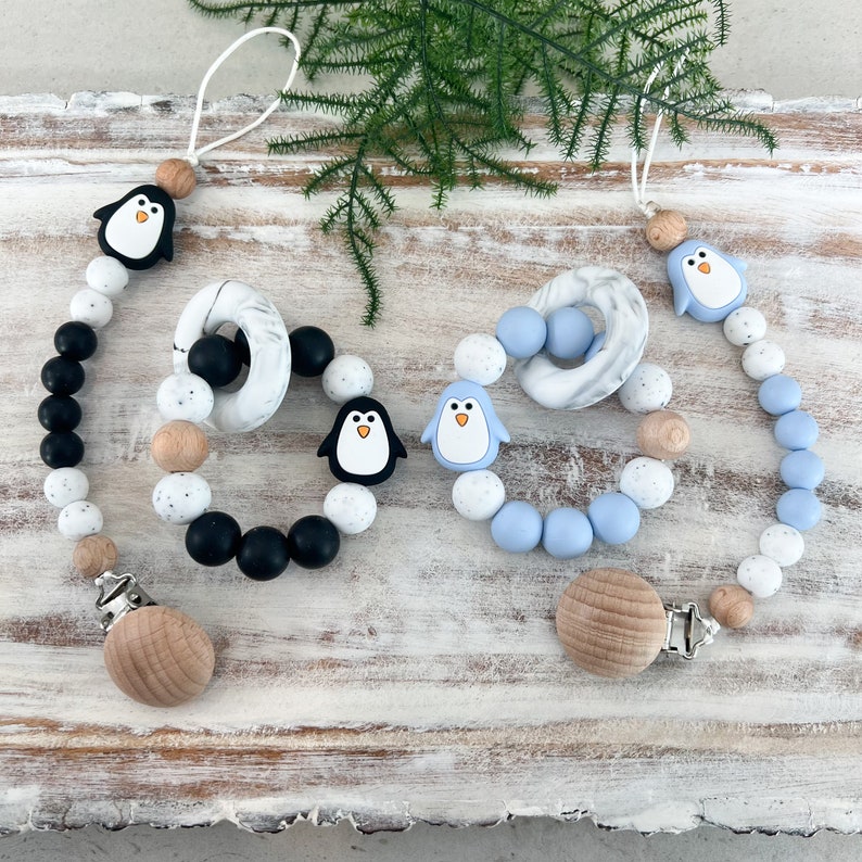 May include: Two baby pacifier clips with penguin designs. One clip features black, white, and tan beads, while the other has blue, white, and tan beads. Each clip has a wooden clasp and a penguin-shaped charm.