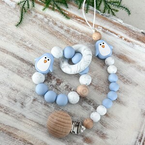 May include: A baby pacifier clip and teether set. The clip features a wooden clip and a string of blue, white, and wooden beads. The teether is a ring with a marble design and two penguin-shaped silicone charms. The set is designed for infants.