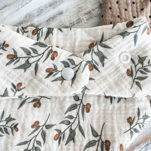K&ouml;nnte beinhalten: Nahaufnahme eines cremefarbenen Baby-L&auml;tzchens mit einem botanischen Druck von Olivenzweigen und braunen Oliven. Das L&auml;tzchen hat einen Druckknopfverschluss und ist aus weichem, strukturiertem Stoff gefertigt. Ein zweites L&auml;tzchen mit Polka-Dots ist teilweise sichtbar.