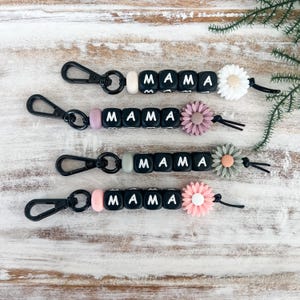 May include: Four keyrings spelling "MAMA" with white letters on black square beads. Each keyring features a different coloured accent bead and a flower charm in white, pink, or grey. The keyrings have black metal clasps.