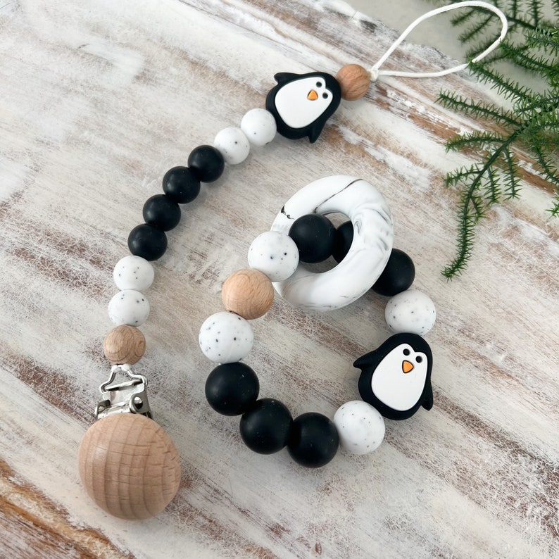 May include: A baby pacifier clip and teether set. The clip features black, white, and wooden beads, a wooden clip, and a penguin charm. The teether is a ring with black, white, and wooden beads and a penguin charm.