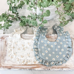 May include: Two baby bibs with ruffled edges. One bib is white with a yellow floral pattern and the other is blue with a white daisy pattern.