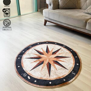 May include: A round, beige and brown rug with a compass design. The compass features a black star with a brown center and the letters N, E, S, and W around the edge. The rug has a black border with small white diamonds.
