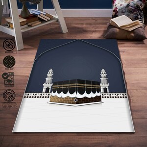 May include: A white and blue prayer rug with a depiction of the Kaaba, a cube-shaped building in Mecca, Saudi Arabia. The rug has a dark blue background with a white border and a white design of the Kaaba in the center. The rug is made of a soft, durable material and is perfect for use in prayer.