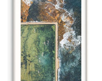 May include: An aerial view of a rectangular ocean pool with green water and several swimmers, bordered by a concrete edge. To the right, dark blue ocean waves crash against jagged, textured orange and brown coastal rock formations.