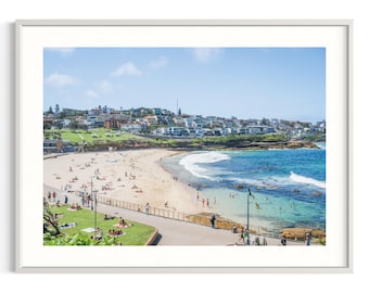 May include: A framed landscape photograph of a coastal beach scene featuring a sandy shore, turquoise ocean waves, and a grassy park area in the foreground. Numerous people are visible relaxing on the sand and swimming in the clear water.