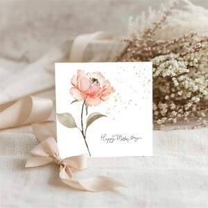 Square Mother’s Day greeting card featuring a soft watercolor pink flower with green leaves and scattered gold speckles, styled on a neutral fabric background with ribbon and dried florals.