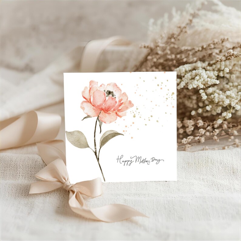 Square Mother’s Day greeting card featuring a soft watercolor pink flower with green leaves and scattered gold speckles, styled on a neutral fabric background with ribbon and dried florals.