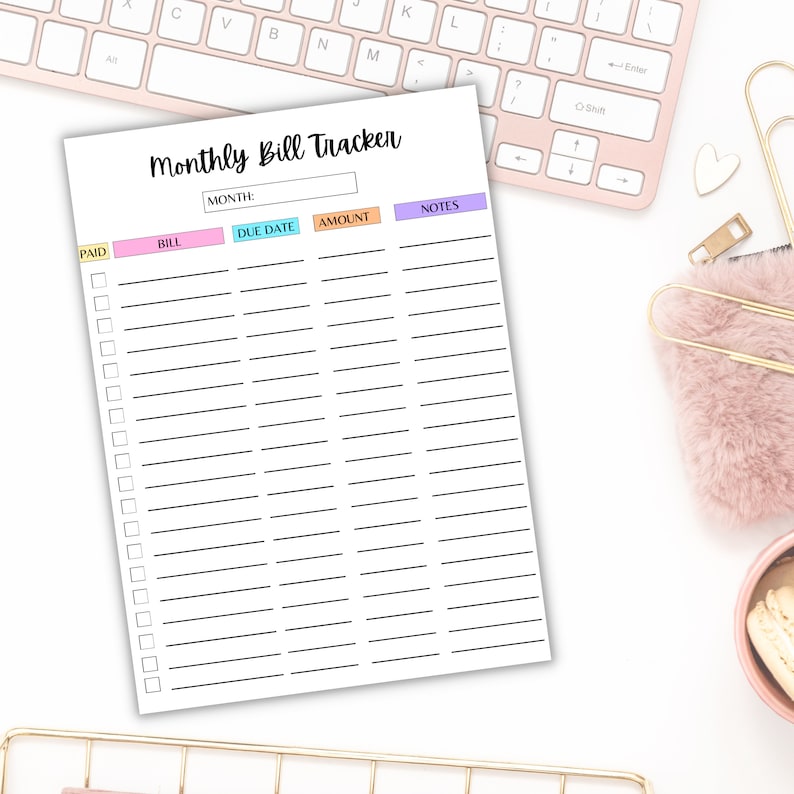 Monthly bill tracker sheet with colorful headers and checkboxes displayed on a soft pink desk with a keyboard, pen, and cozy accessories.