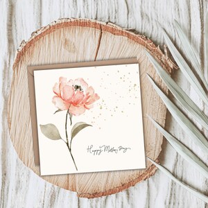 A square floral greeting card featuring a watercolor pink flower, styled on a wood slice with greenery accents.