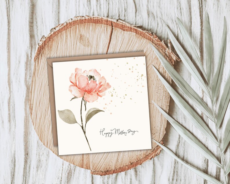 A square floral greeting card featuring a watercolor pink flower, styled on a wood slice with greenery accents.
