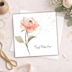 Square Mother’s Day card with watercolor blush pink flower and gold speckles styled with envelope, ribbon, and gold scissors on soft neutral background.