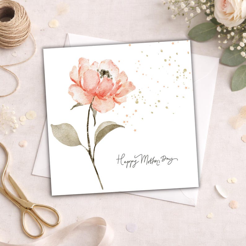 Square Mother’s Day card with watercolor blush pink flower and gold speckles styled with envelope, ribbon, and gold scissors on soft neutral background.