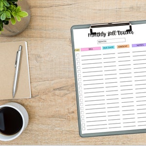 Monthly bill tracker sheet on a clipboard with colorful column headers, displayed on a wooden desk beside a notebook, pen, coffee cup, and a small plant.