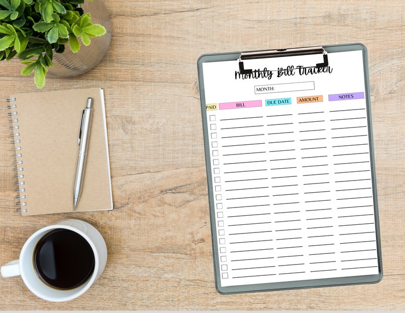 Monthly bill tracker sheet on a clipboard with colorful column headers, displayed on a wooden desk beside a notebook, pen, coffee cup, and a small plant.