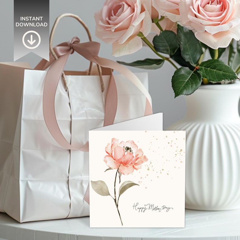 Square floral greeting card with a watercolor pink flower design displayed in front of a gift bag and vase of roses on a styled tabletop.