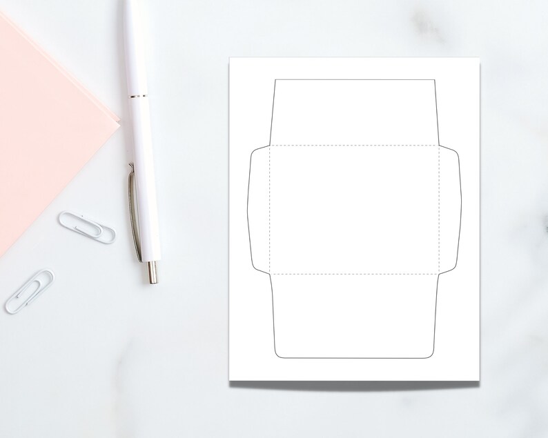 Envelope template sheet displayed on a marble surface with a white pen and paperclips, showing the solid and dotted cut-and-fold outline.