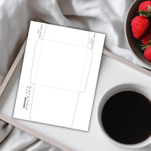 Square 5x5 envelope template shown on a tray beside a cup of coffee and a bowl of strawberries, with cut and fold lines visible on the page.